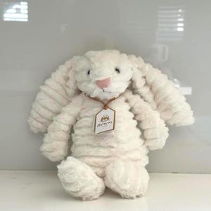 Puede incluir: Un conejito de peluche color crema con largas orejas caídas y una nariz rosa. El peluche tiene un aspecto texturizado, similar a la pana, y una pequeña etiqueta que dice "Jellycat".