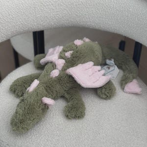 May include: A plush green alligator toy with pink accents on its back, feet, and wings. The stuffed animal is lying on a white chair. A small tag is attached to the toy.