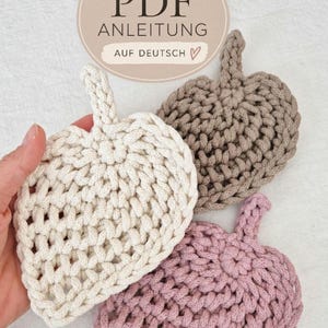 May include: Three crocheted leaf-shaped coasters in cream, taupe, and dusty pink. Each coaster features a central stem and a textured, woven stitch pattern. A circular label at the top displays the text PDF ANLEITUNG AUF DEUTSCH.