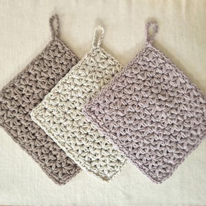 Könnte beinhalten: Drei handgefertigte Topflappen aus Häkelarbeit in Grau-, Off-White- und Lavendeltönen. Jeder quadratische Topflappen hat ein strukturiertes, sternförmiges Muster und eine Schlaufe zum Aufhängen. Küchenzubehör.