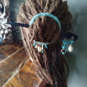 以下が含まれることがあります： ターコイズのヘアバンドと黒いヘアスティックでスタイリングされた茶色のドレッドヘアのクローズアップ。ヘアアクセサリーには、小さなターコイズビーズと白い貝殻のアクセントが施され、ボヘミアンスタイルを演出しています。