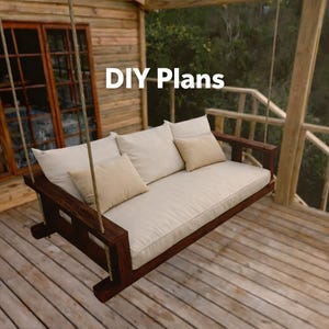 May include: A dark brown wooden porch swing with a rectangular frame, suspended by thick ropes. The swing features a large off-white seat cushion, two matching back cushions, and two smaller beige accent pillows. The text DIY Plans is visible.