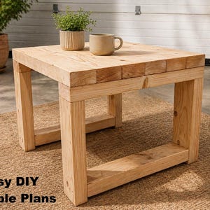 Puede incluir: Una mesa cuadrada de madera clara con un acabado natural. La mesa tiene un diseño simple y macizo con patas gruesas y una parte superior resistente. Una pequeña planta en maceta y una taza beige están sobre la mesa. El texto "Easy DIY Table Plans" está en la parte inferior.
