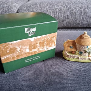 Könnte beinhalten: Eine Miniatur-Cottage-Figur mit Strohdach und Steinmauern, neben einer grünen Schachtel. Auf der Schachtel stehen die Worte "Lilliput Lane" und "Handmade in England Treasured Worldwide". Das Cottage hat einen kleinen Garten.