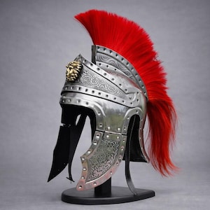 Op de afbeelding: Een zilverkleurige Romeinse helm met een felrode pluim. De helm heeft ingewikkelde reliëfontwerpen en een goudkleurig leeuwenkop-embleem. Het wordt tentoongesteld op een zwarte standaard, wat het historische ontwerp benadrukt.
