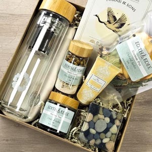 May include: A gift box filled with self-care items. It includes a glass tea infuser bottle with a bamboo lid, jars of bath salts and tea, a yellow lotion tube, a bar of soap, and a package of dried fruit. The box also contains a card with a crane illustration.