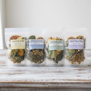 May include: Four clear pouches of simmer pot blends, each with a label reading "EDITH & EDEE Simmer Pot." The pouches contain dried herbs and citrus slices. The labels are in pastel colors, with text indicating the blend names.