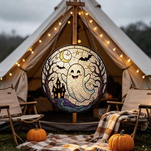 Puede incluir: Un atrapasoles circular de vidrieras que presenta un fantasma blanco central con cara sonriente, una casa encantada negra, una luna creciente amarilla, estrellas y murciélagos negros. La pieza está enmarcada en metal negro y cuelga de una cadena.