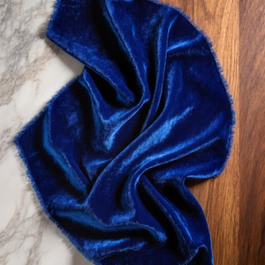 Pode incluir: Um tecido de veludo azul vibrante com uma textura macia e brilhante. O tecido é drapeado, mostrando sua cor rica e brilho sutil. As bordas são desfiadas, adicionando um toque rústico. O fundo é uma mistura de mármore e madeira.