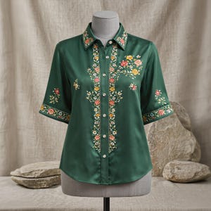 Puede incluir: Una camisa verde oscuro con mangas cortas, con bordados florales en rosa, amarillo y blanco. El bordado está en el cuello, en la parte delantera y en los puños de las mangas. La camisa se exhibe en un maniquí.