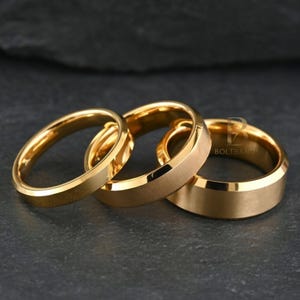May include: Three gold-colored rings with varying designs. One ring has a smooth, polished surface, while the others have a brushed or matte finish. The rings are displayed on a dark, textured surface.