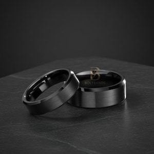 May include: Two black rings with a brushed metal finish. One ring is angled towards the viewer, while the other is positioned behind it. The rings are on a dark surface.