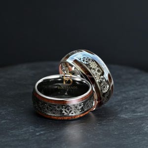 May include: Two silver rings with a gear design and wood inlays. The rings have a dark interior and a polished exterior. The wood inlays are a reddish-brown color. The rings are from BoltBands.