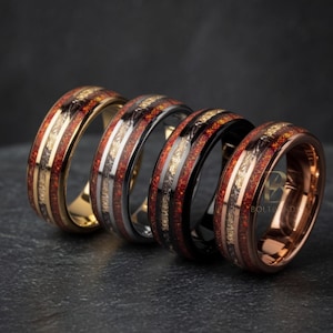 Op de afbeelding: Vier ringen met een uniek ontwerp. Elke ring heeft een band van rood en goudkleurig materiaal, met een metalen streep. De ringen zijn in goud, zilver, zwart en roségoud. De ringen liggen op een donkere ondergrond.