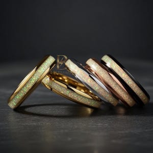 May include: A collection of five rings with opal inlays. The rings feature gold, silver, and rose gold bands, with the opal inlays displaying a mix of white, green, and iridescent colors. The rings are displayed on a dark surface.