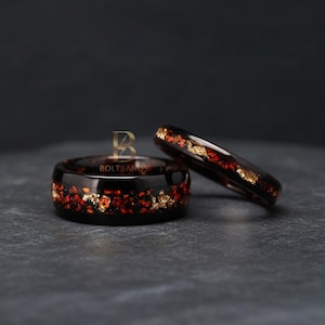 May include: Two black resin rings with embedded red and gold flakes. The rings are displayed on a dark surface. The rings have a glossy finish and a unique design. These rings could be wedding bands or fashion accessories.