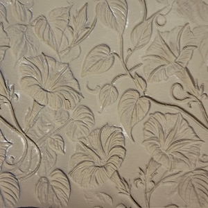 May include: A close-up of a beige surface with a floral relief pattern. The design features morning glory flowers and leaves, with intricate details and shadows creating a textured appearance. The relief work is consistent throughout the image.