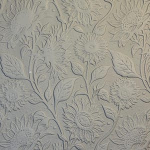 May include: A white surface featuring a repeating embossed pattern of sunflowers with detailed petals, central seed heads, and winding stems with leaves. The background has a subtle stippled texture that contrasts with the raised floral design.