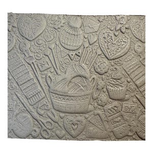 May include: A grey relief sculpture with a textured surface, depicting craft-related items. The design includes a basket of yarn, knitting needles, scissors, hearts, a cupcake, and a knitted hat. The theme suggests handmade crafts and hobbies.