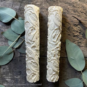 May include: Two cream-coloured, cylindrical rolling pins featuring embossed bird and floral patterns. The text "77mm Embossed & Debossed" is at the bottom. The pins are displayed on a wooden surface with eucalyptus leaves.