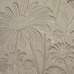 May include: A close-up of a beige, textured surface with an embossed floral design. The design features detailed daisies, buds, and leaves. The relief pattern creates a three-dimensional effect, showcasing the intricate details of the flowers.