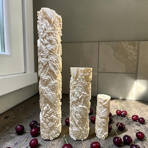 May include: Three cream-coloured, carved decorative pillars of varying heights, each intricately detailed with floral and leaf patterns. The pillars stand on a wooden surface, surrounded by fresh, ripe cherries. The tallest pillar is approximately 25 cm tall.