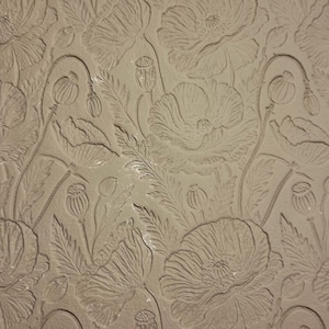 May include: A close-up of a beige surface with an embossed floral pattern. The design features detailed outlines of poppies, leaves, and seed pods. The relief creates a textured, three-dimensional effect across the surface.