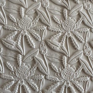 May include: A light grey, matte-finish surface featuring a repeating, embossed pattern of protea flowers and elongated leaves. The raised, three-dimensional design creates a textured, monochromatic floral motif across the entire flat surface.