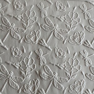 May include: A light beige fabric featuring an embossed, repeating pattern of dragonflies and five-petalled flowers. The design is monochromatic, with the raised, textured motifs standing out against the flat, smooth background of the textile.