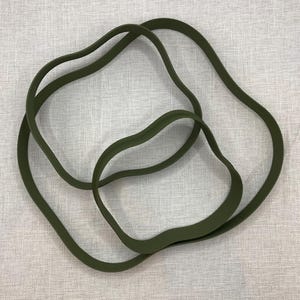 May include: Three olive green, wavy rubber bands are intertwined, forming an abstract design. The bands have a smooth texture and are set against a neutral, textured backdrop. The bands' organic shapes suggest flexibility and movement.