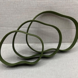 May include: Three olive green, abstract, wavy-shaped objects are intertwined on a light grey surface. The objects have a smooth, matte finish and vary in size, creating a layered effect. The shapes appear to be made of a flexible material.