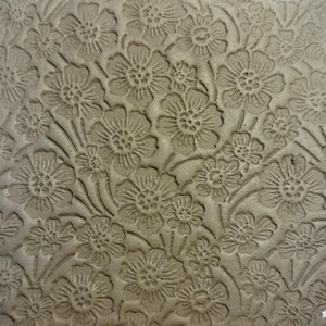 May include: Close-up of a textured, neutral-toned surface with an embossed floral pattern. The design features detailed flowers and leaves, creating a repeating, natural motif. The raised elements cast subtle shadows.