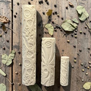 May include: Three off-white rolling pins with embossed floral and botanical designs. The pins are of varying lengths, the longest displaying sunflowers and intricate patterns. The background is a wooden surface with scattered leaves and seeds.