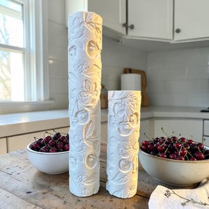 May include: Two white ceramic vases with a cherry blossom design. The vases are cylindrical, one tall and one short, and are placed on a wooden table. Bowls of fresh cherries are on either side of the vases. The background is a kitchen setting.