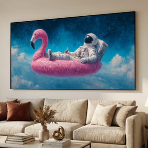 May include: A large framed canvas art print depicts an astronaut relaxing on a pink flamingo pool float in a dreamy, cloud-filled sky. The astronaut is wearing a white spacesuit and holding a beverage. The background features a dark blue, starry sky.
