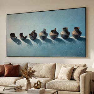 May include: A large framed painting depicts a row of abstract, dark-colored pottery against a light blue background. The pottery has a rough texture and varying shapes, with some pieces appearing broken. The artwork is displayed above a beige sofa.