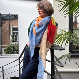 Może przedstawiać: Kolorowa, dziergana chusta w odcieniach niebieskiego, pomarańczowego, czerwonego, żółtego i beżowego. Chusta jest zarzucona na ramiona. Osoba ma na sobie zielony top i ciemnoniebieskie dżinsy. Tło obejmuje schody i okno.