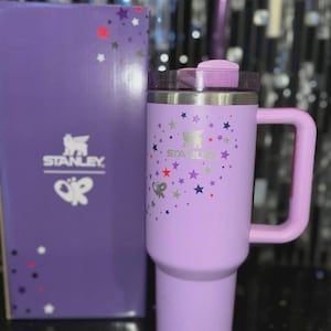 May include: A lavender Stanley travel mug with a handle, lid, and the Stanley logo. The mug is decorated with colorful star graphics. A matching lavender box with the Stanley logo and star graphics is next to the mug.
