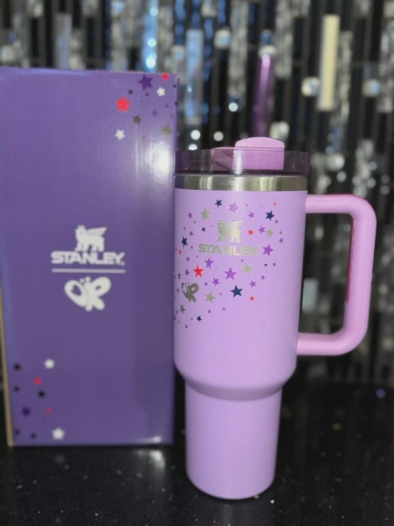 May include: A lavender Stanley travel mug with a handle, lid, and the Stanley logo. The mug is decorated with colorful star graphics. A matching lavender box with the Stanley logo and star graphics is next to the mug.