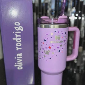 May include: A lavender Stanley tumbler with a handle, lid, and straw, next to a matching box. The tumbler features a star pattern and the Stanley logo. The box has the name "olivia rodrigo" printed on it.