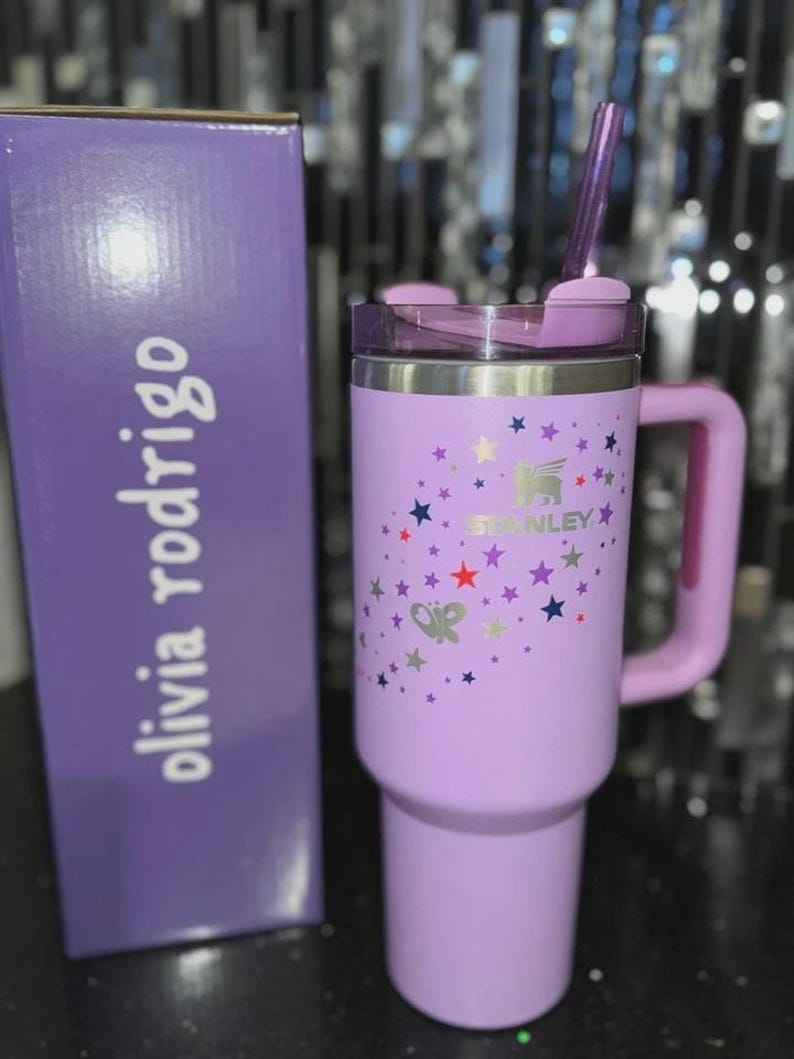 May include: A lavender Stanley tumbler with a handle, lid, and straw, next to a matching box. The tumbler features a star pattern and the Stanley logo. The box has the name "olivia rodrigo" printed on it.