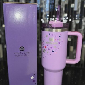 May include: A lavender insulated tumbler with a handle and a lid, next to a matching box. The tumbler features a butterfly and star design with the brand name "STANLEY". The box has social media handles printed on it.