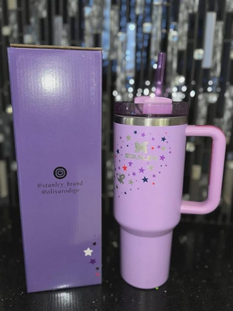 May include: A lavender insulated tumbler with a handle and a lid, next to a matching box. The tumbler features a butterfly and star design with the brand name "STANLEY". The box has social media handles printed on it.