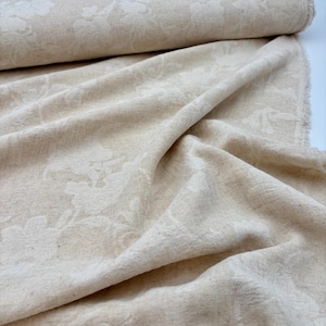 Può includere: Un rotolo di tessuto beige chiaro caratterizzato da un motivo floreale tono su tono intrecciato nel materiale. Il tessuto presenta una trama visibile ed è drappeggiato in morbide pieghe, mostrando l'intricato disegno botanico sulla superficie.