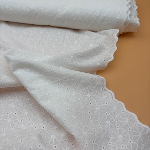Op de afbeelding: Witte eyelet stof met een bloemmotief en geschulpte randen. De stof is gevouwen en opgerold, wat de textuur laat zien. Geschikt voor diverse naai- en knutselprojecten. Afmetingen in centimeters.