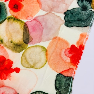 Può includere: Un tessuto con un motivo floreale ad acquerello. Il design presenta grandi forme sovrapposte in tonalità di rosso, arancione, rosa, verde e beige su sfondo bianco. I bordi del tessuto sono visibili.