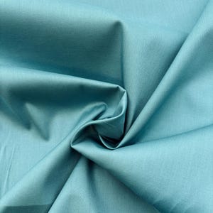 Könnte beinhalten: Ein einfarbiger, blaugrüner Stoff mit einer glatten, matten Oberfläche. Das Material ist in weichen Falten drapiert, die tiefe Schatten werfen und die gleichmäßige Farbe sowie die feine, gewebte Struktur des Textils deutlich hervorheben.