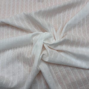 Puede incluir: Tela blanca con un patrón texturizado de puntos y rayas verticales. La tela está doblada y drapeada, creando sombras y reflejos. El material parece suave y ligero, adecuado para ropa o decoración del hogar.