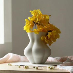 Puede incluir: Un jarrón de cerámica blanca con una forma curva única, que contiene un vibrante arreglo de flores amarillas brillantes. El jarrón está sobre una superficie de madera, con un paño rosa suave en el fondo. Las flores añaden un toque de color a los tonos neutros.