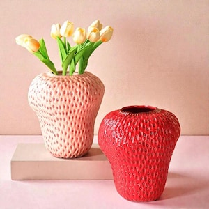 Op de afbeelding: Twee keramische aardbeivormige vazen. Eén is lichtroze en bevat gele tulpen. De andere is felrood. Beide vazen hebben een gestructureerd oppervlak dat aardbeienzaden nabootst. De vazen staan op een roze ondergrond.